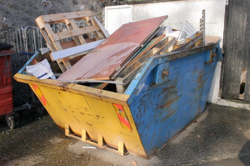 A large industrial metal skip painted yellow on the front and blue on the sides, situated on a paved surface outdoors, filled with various discarded construction and renovation materials. The contents include wooden pallets, pieces of particle board, and metal fixtures, all stacked haphazardly with some leaning against the sides of the skip. The wooden pallets have a weathered appearance with visible grain and some broken or missing slats, while the particle board exhibits a smooth, light brown finish. The metal fixtures, including a partially visible metal grate, show signs of rust and wear. The skip is positioned against a concrete wall on the right and a dark wooden fence on the left, with a portion of a neighbouring structure visible behind. The surroundings appear to be an industrial or commercial back area, typical of waste collection sites in the locality near Mottingham, offering a clear view of waste materials ready for removal by a professional rubbish clearance service like Commercial Waste Mottingham.