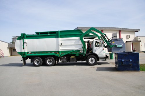 A large commercial waste collection truck operated by Commercial Waste Mottingham is parked on a paved driveway in a residential or commercial area. The truck has a white cab with a green and white loading container, featuring a sturdy metal construction with visible rivets and reinforced edges. The vehicle's hydraulic lifting arm is positioned at the rear, suggesting it is in the process of collecting or depositing rubbish. To the right of the truck, there are two large, open waste containers—one dark blue and the other grey—placed on the ground, ready for waste disposal. The background shows a modern, light-colored building with large windows and a flat roof, situated in an area with suburban or urban development typical of the vicinity of Mottingham in London. The scene is illuminated by natural daylight, with clear skies overhead, highlighting the industrial and functional nature of the waste collection operation carried out by Commercial Waste Mottingham in this localized setting.