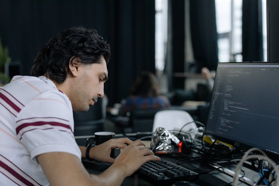 A young man with dark, wavy hair is seated at a desk in an office environment, working on a computer with a large monitor displaying lines of code. He is wearing a short-sleeved, white shirt with maro