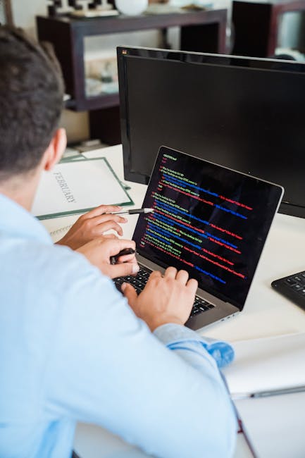 A young man with dark, wavy hair is seated at a desk in an office environment, working on a computer with a large monitor displaying lines of code. He is wearing a short-sleeved, white shirt with maro